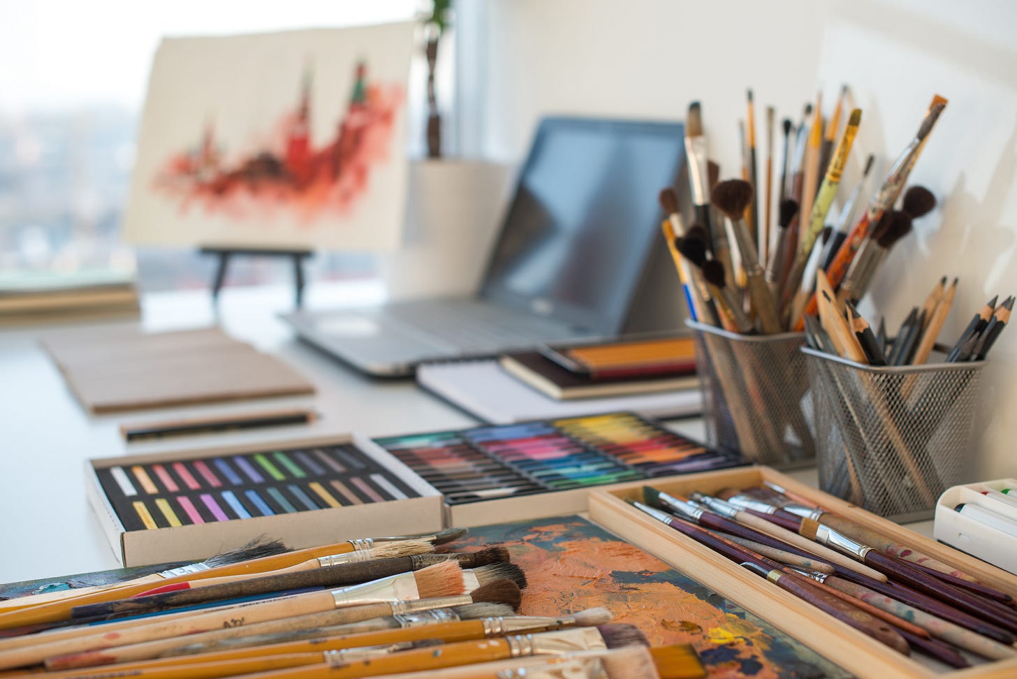 paintbrushes, pencils, pastels, a laptop, and various other art supplies on a desk