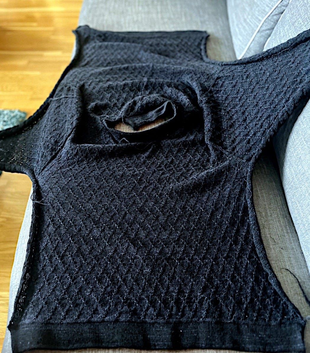 A black machine-knit sweater laid flat on a grey sofa, showing a diamond stitch texture across the body. The two completed sleeves extend out to either side. A black machine-knit sweater laid flat on a grey sofa, showing a diamond stitch texture across the body. The two completed sleeves extend out to either side.
