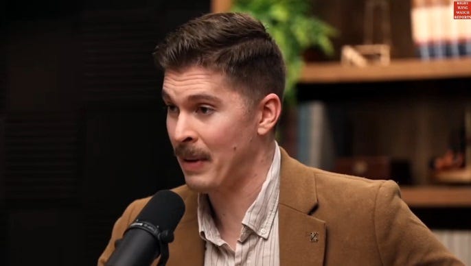 A mustached white man in a brown suit with no tie speaks into a microphone on a podcast set, with a bookshelf in the background. Only a few books are on the shelf, which also holds a potted plant, a small lidded box, and some unidentifiable out of focus stuff