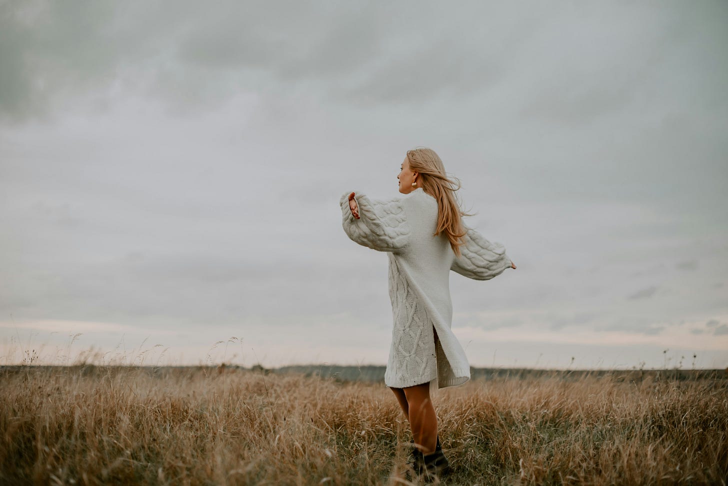 A person in a long, light-colored sweater stands in a grassy field with arms spread, facing cloudy skies representing freedom. A person in a long, light-colored sweater stands in a grassy field with arms spread, facing cloudy skies representing freedom.