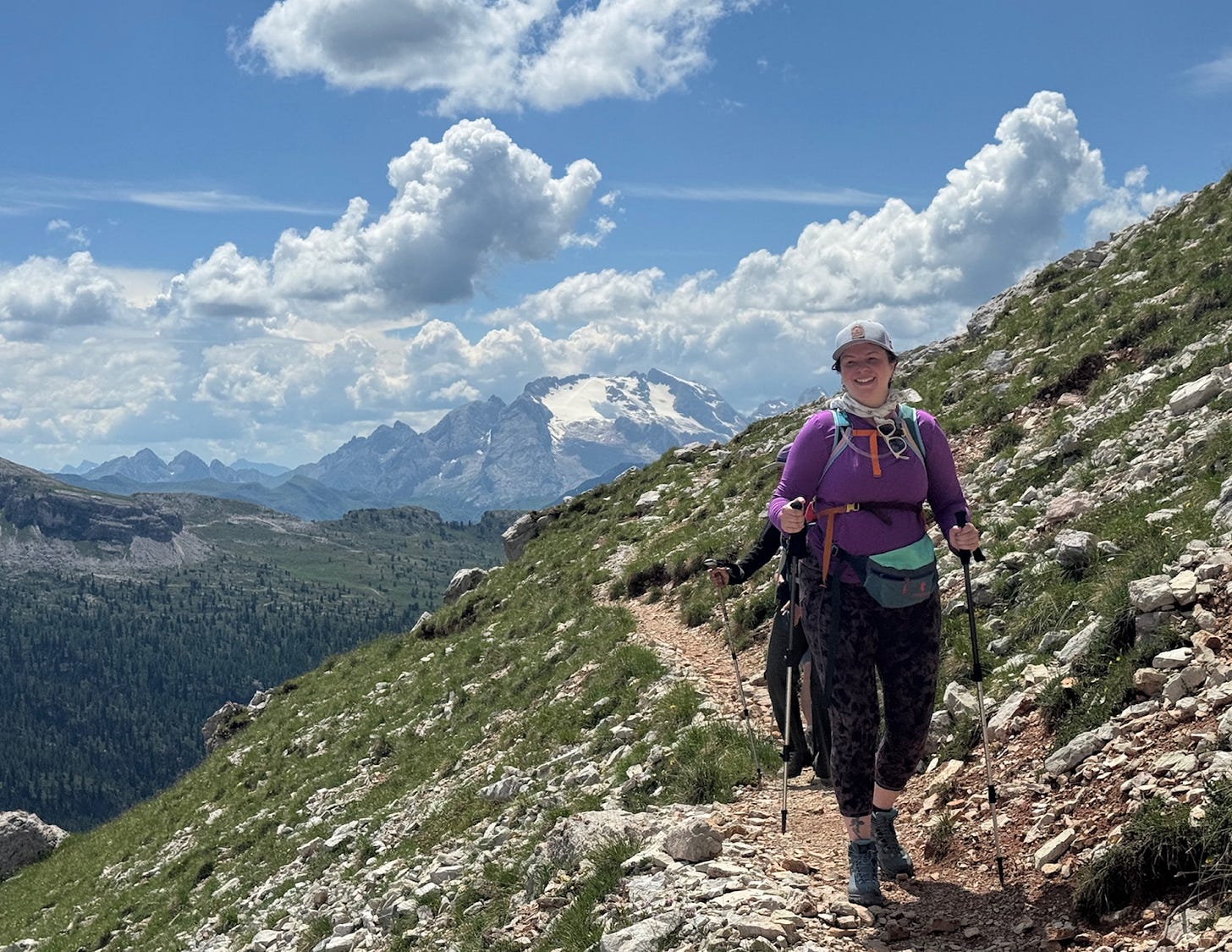 Amber walks on a narrow trail on the side of a mountain. She uses trekking poles. The sky is a bright blue with big, puffy clouds. Other mountains can be seen in the distance.
