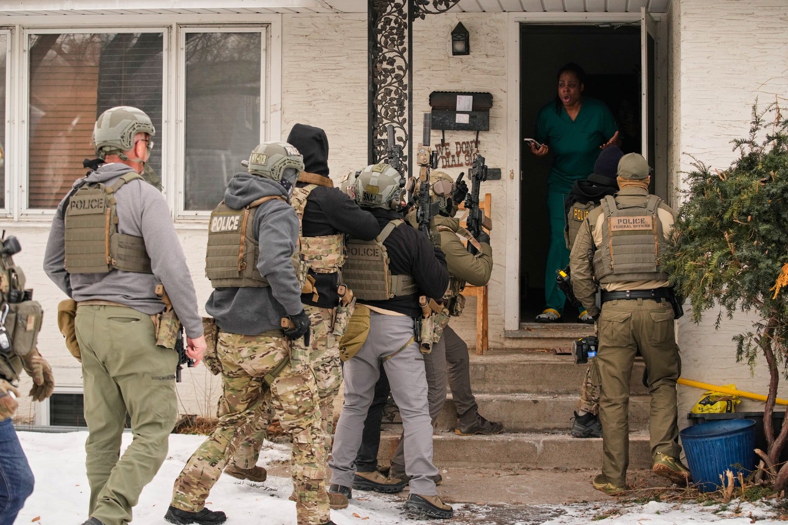 At least eight armed federal officers stand outside the open door of a house as a person inside gestures while speaking to them.