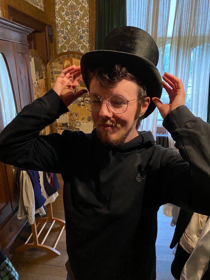 Two photos of me, a white trans masc with round glasses and dark blonde/light brown hair – one in the Abbey gardens, framed by a bare topiary tree archway, the second inside trying on a black top hat