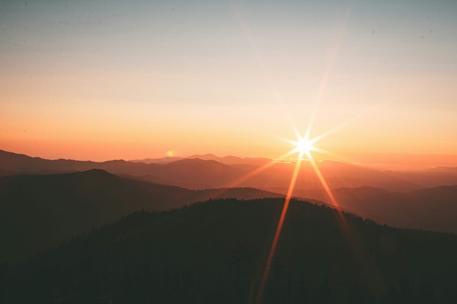 The sun rises or sets above the tops of some forested mountains. The colors of the photo blur together and are mostly orange/red. The sun rises or sets above the tops of some forested mountains. The colors of the photo blur together and are mostly orange/red.