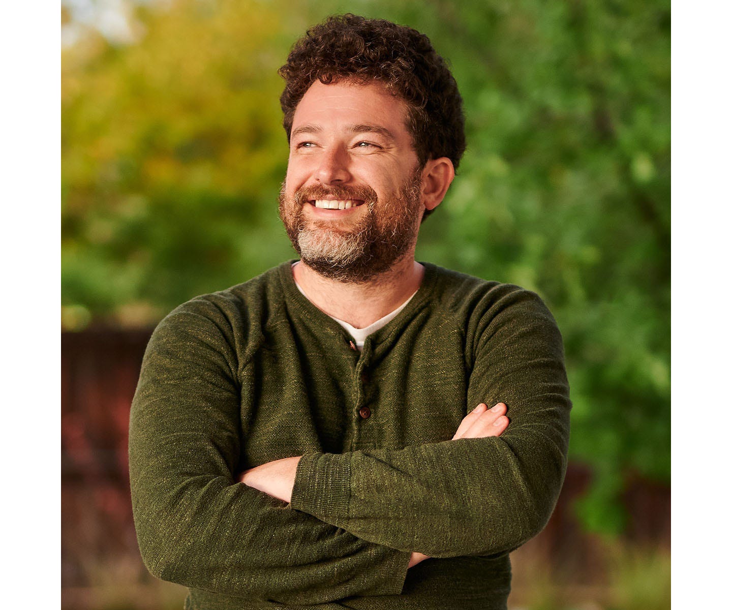 A young, bearded white man stands smiling and look off to one side with his arms crossed.