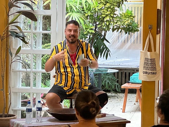 Na primeira imagem, estou sentado em uma cadeira alta, falando e gesticulando com um jardim de inverno ao fundo. Visto camisa listrada amarela e preta. Sou um homem de pele bege clara, olhos, cabelos, barba e bigode escuros e gordo. A segunda imagem é uma foto semelhante à primeira mas com o plano aberto, enquadrando também a sala da casa e os espectadores da mesa, ao menos umas 8 pessoas.