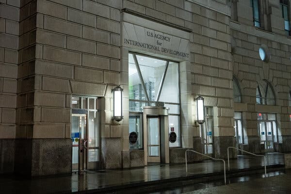 The exterior of a white stone building with a sign above the revolving door that says “U.S. AGENCY for INTERNATIONAL DEVELOPMENT,” seen at night.