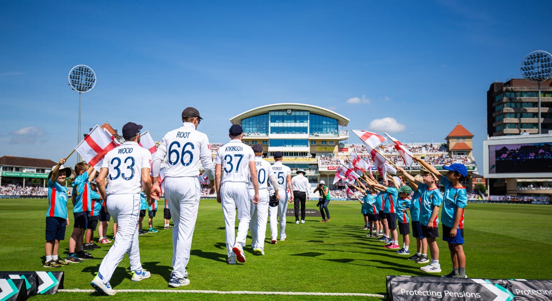 A cricket team in Test whites walking out onto a green cricket pitch on a sunny day, with children in blue shirts holding up England flags around them