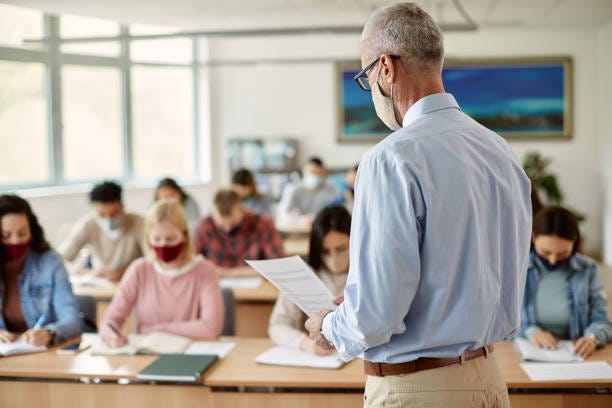 Rear view of teacher wearing face mask and giving lecture to group of students in the classroom. Rear view of teacher wearing face mask and giving lecture to group of students in the classroom.
