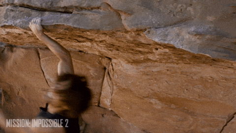 Scena d'azione dal film Mission: Impossible 2. Un uomo con i capelli lunghi (interpretato da Tom Cruise) si aggrappa con una sola mano a una parete rocciosa a strapiombo, oscillando leggermente mentre cerca di mantenere la presa.