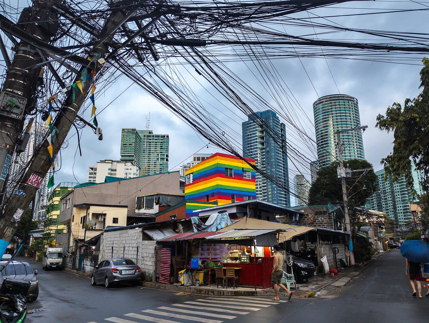 A street corner in a Manila neighborhood. Simple 2 and 3 story concrete and wood buildings, a little stor on the corner, some laundry hanging from a window. Two utility poles are in the upper left. They are overflowing with wiring, to the point of leaning precariously over. In the distance, some highrises. In the center of the photo, a multi-story building painted with bright horizontal stripes of the rainbow.