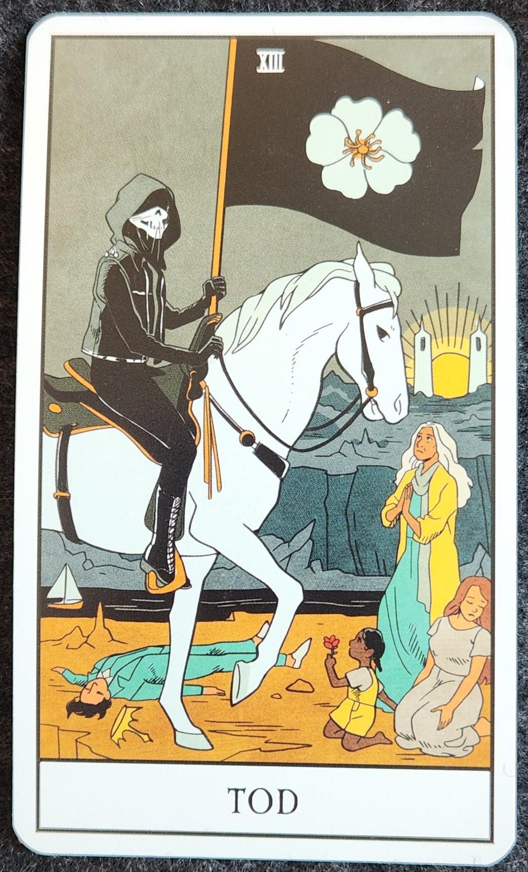 A tarot card labelled XIII Tod. A skeletal figure in a black hooded jacket is riding a white horse. They are carrying a black flag with a white 5-petaled flower on it. On the ground surrounding them are 4 people. One person lays dead on their back, a golden crown fallen from their head. A small child kneels in front of the horse, holding up a red flower. A young woman kneels behind the child with her eyes closed and face slightly turned away. An older woman stands in front of the horse, looking up at the rider with her hands pressed into a prayer position. In the background there is a black river and a grey rocky landscape. Beyond, a structure with two white towers frame the sun rising.