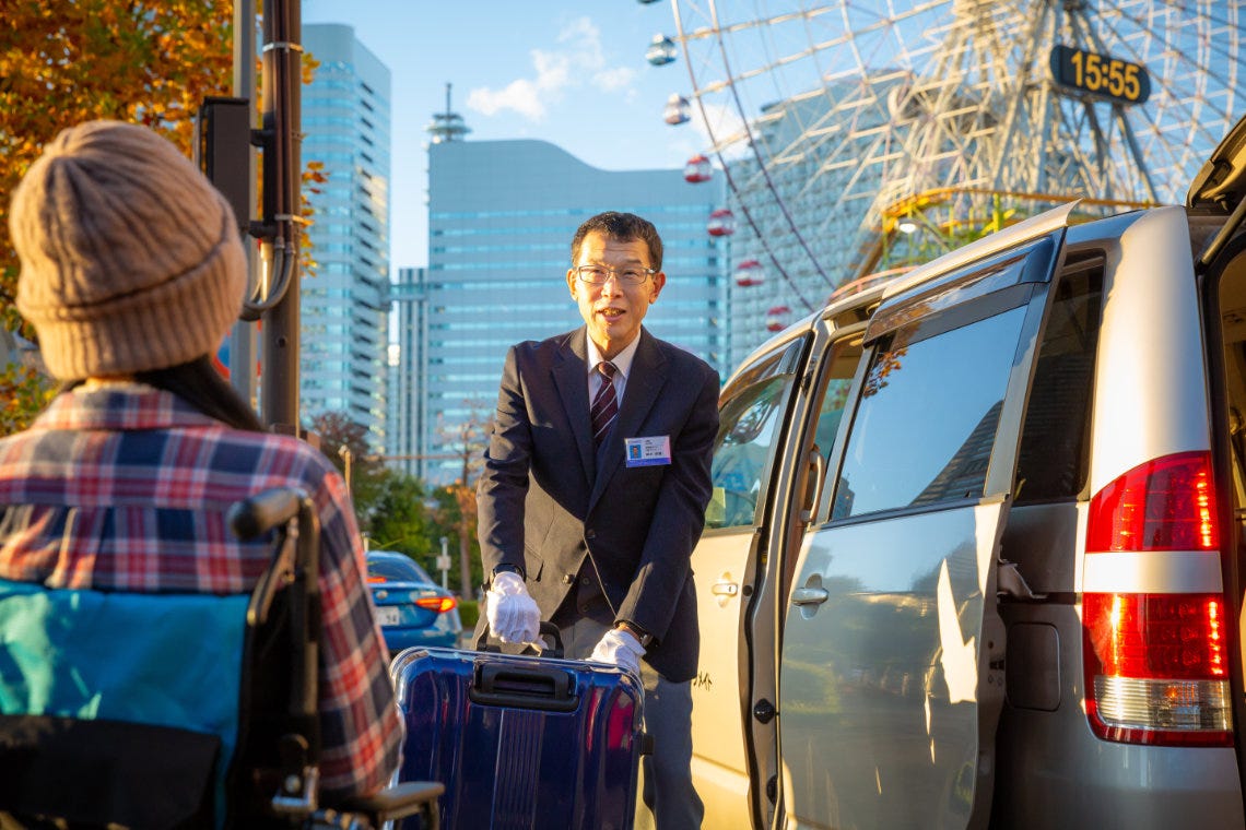 Yoshio Suzuki in a dark suit and white gloves lifting a blue suitcase into his van, a wheelchair user in a knit hat in the foreground, with the Cosmo Clock Ferris wheel and Minato Mirai skyline behind.