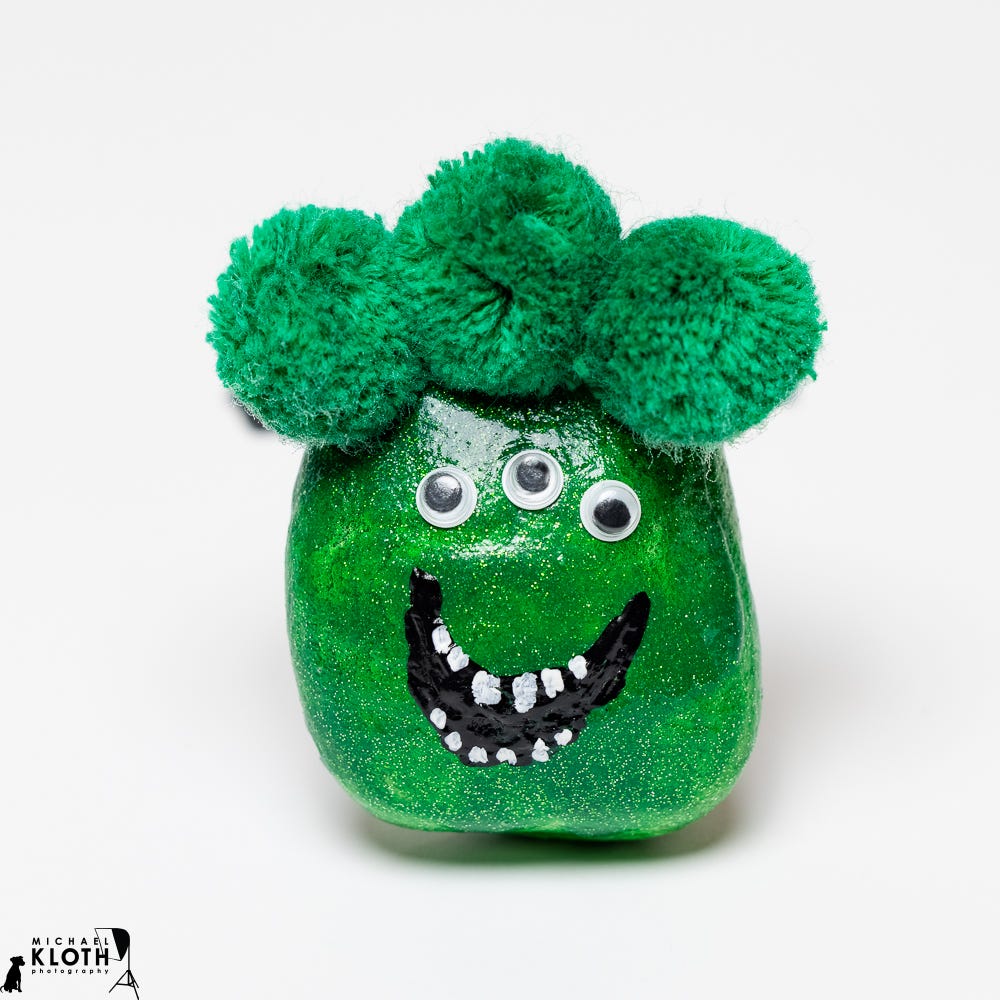 A green rock 'monster' with three puff balls of 'hair' and three googly eyes photographed in a white studio background. A green rock 'monster' with three puff balls of 'hair' and three googly eyes photographed in a white studio background.