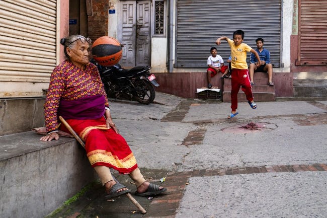 Des enfants jouent au ballon, mais celui-ci est parti en direction d'une grand-mère assise à côté. La photo est prise juste avant impact, la grand-mère n'est aps encore touchée mais l'enfant affiche déjà une mine déconfite