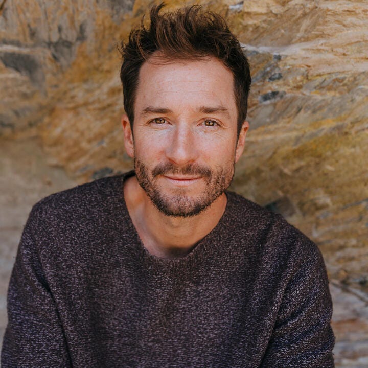 Author photo, man in gray sweater, brown hair, sitting in front of rock wall