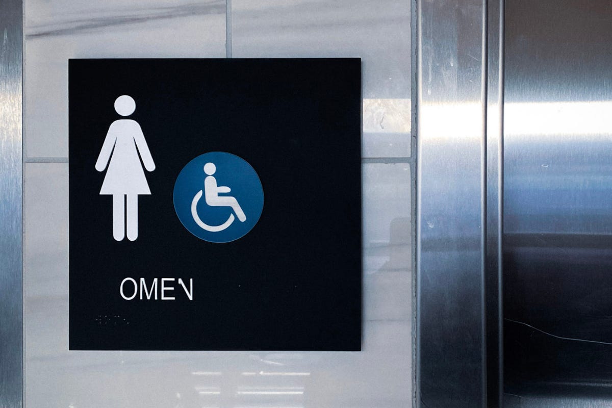 A black, square women's restroom sign with an accessibility symbol, mounted on a white marble-tiled wall. The word "OMEN" is printed in white capital letters below the symbols. A black, square women's restroom sign with an accessibility symbol, mounted on a white marble-tiled wall. The word "OMEN" is printed in white capital letters below the symbols.