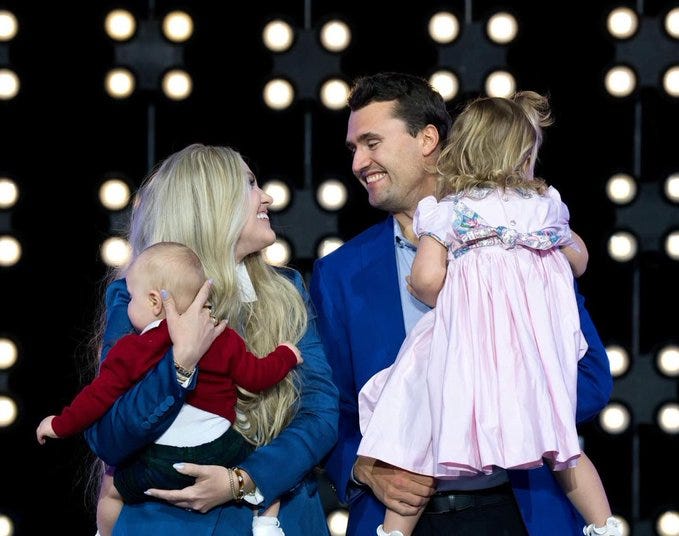 Charlie Kirk stands smiling, wearing a blue suit, holding a young girl in a pink dress with a floral bow. A woman with long blonde hair, wearing a blue jacket, holds a baby in a red outfit, also smiling. The background features a black wall with bright circular lights.