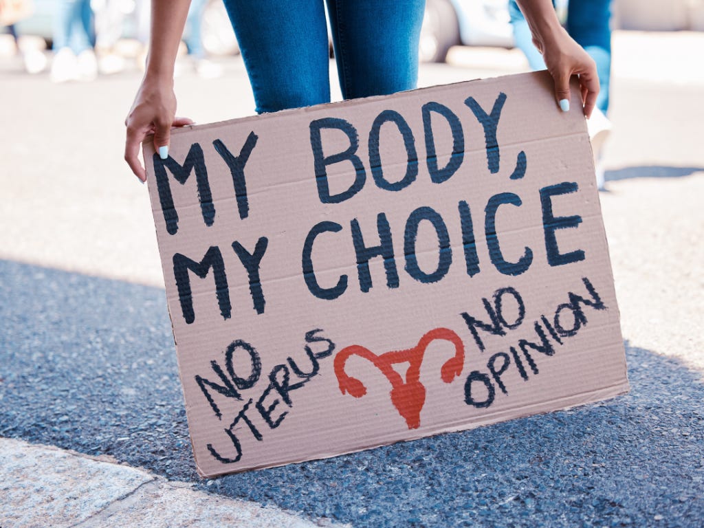 Persoon houdt een kartonnen bord vast met de tekst “My Body, My Choice” en “No uterus, no opinion”.