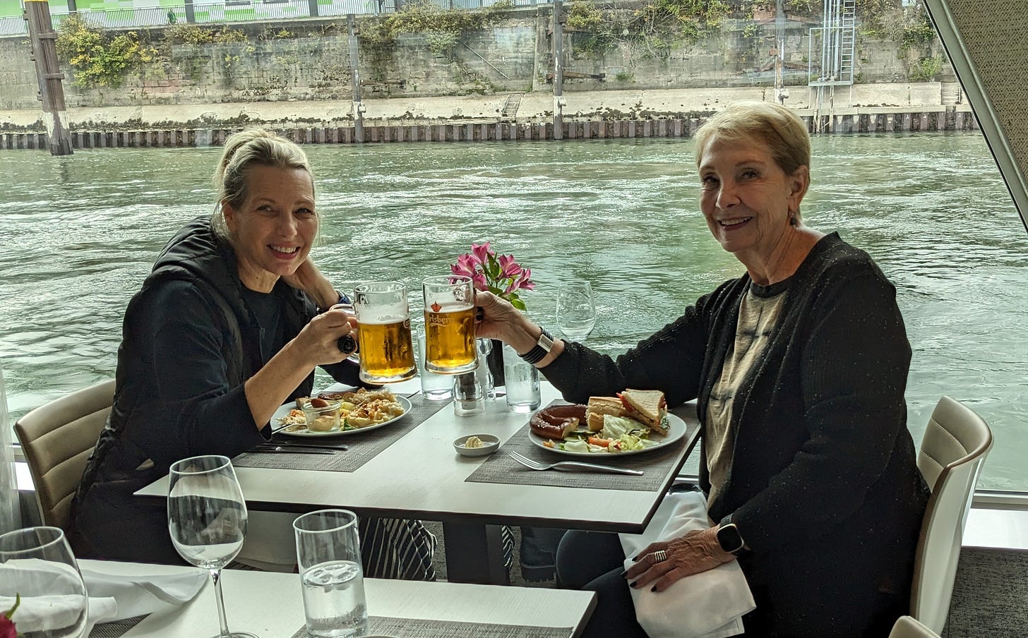 Two white women with bond hair, a mother and daughter, are dining with the river just beyond. They are holding mugs of beers in a toast. 