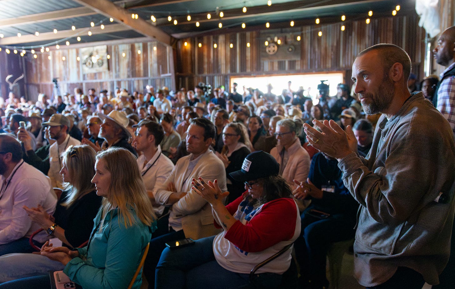 Crowd of attendees sits in a rustic barn under string lights, clapping during a session at the regenerative conference.