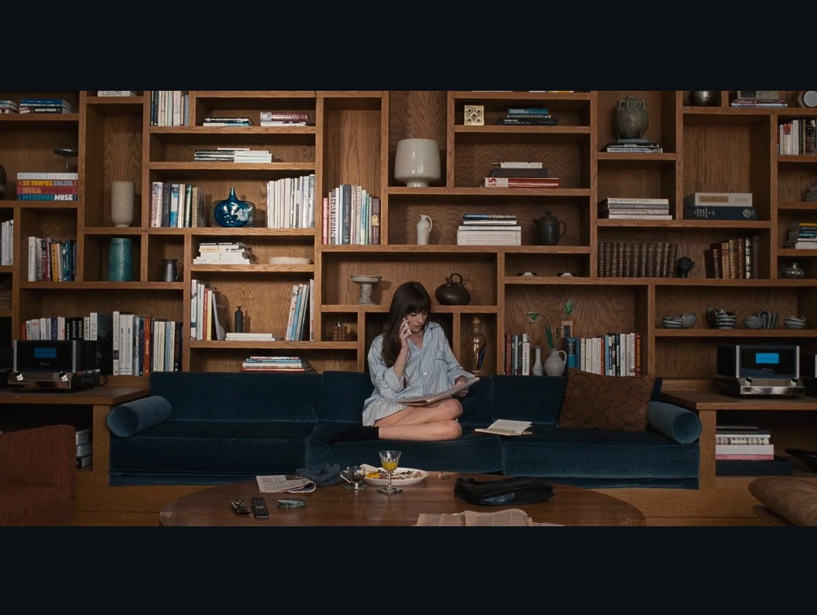 Dakota Johnson sitting on a couch in middle of frame; in front of her a luxurious table; behind her bookshelves with a range of different shapes and orientations of shelves, containing books and expensive tschotskes. Dakota Johnson sitting on a couch in middle of frame; in front of her a luxurious table; behind her bookshelves with a range of different shapes and orientations of shelves, containing books and expensive tschotskes.