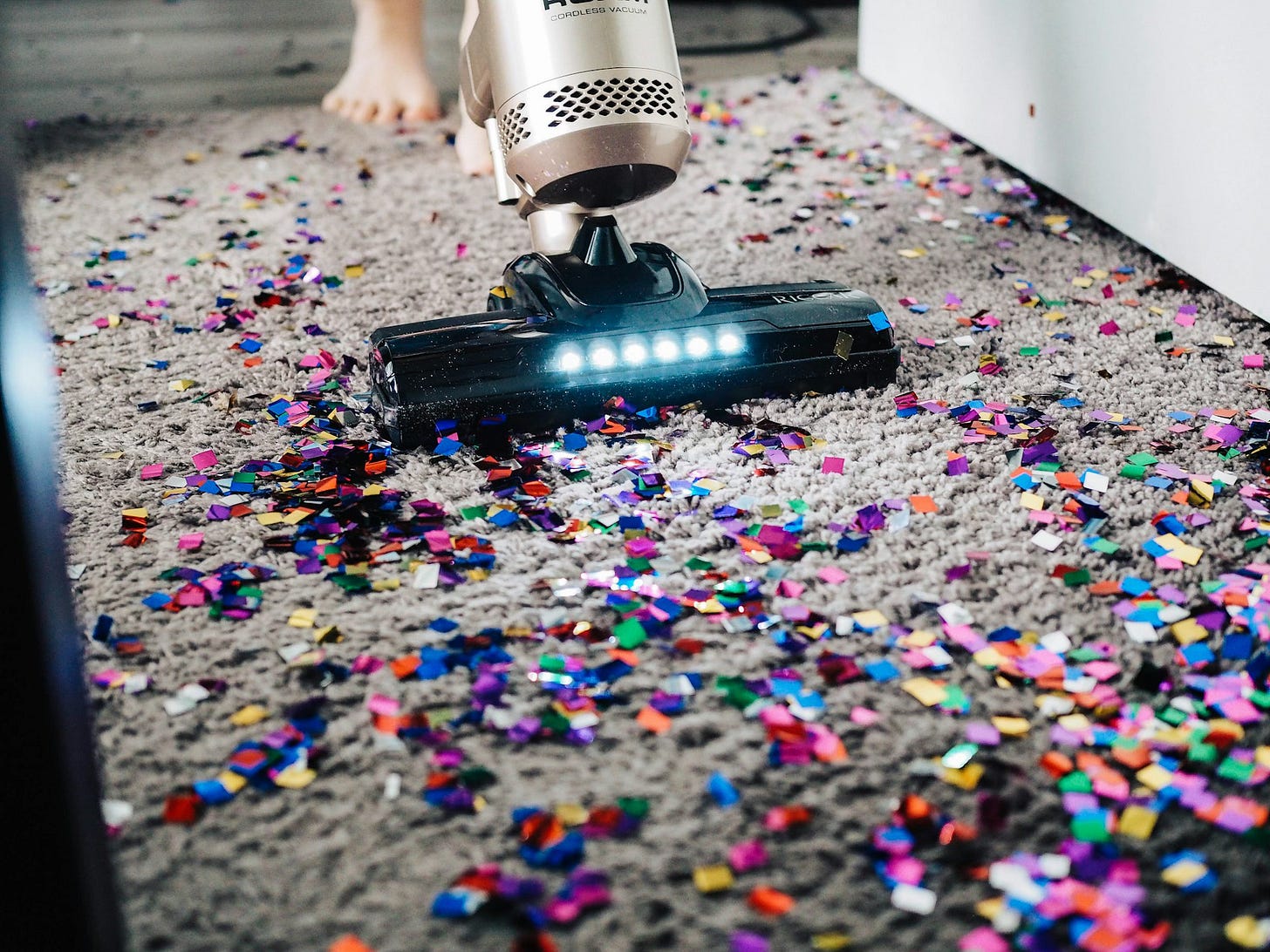 A person with bare feet vacuums up multi-colored  paper confetti off a strip of taupe carpeting.