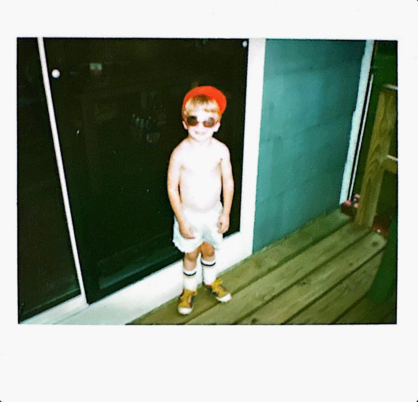 A young child wearing oversized sunglasses, a ballcap, and cowboy boots, standing on a wooden deck. Half costume, half armor.