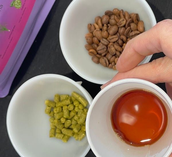 photo of coffee in foreground, light amber color, behind in background are two bowls with the beans and hop pellets