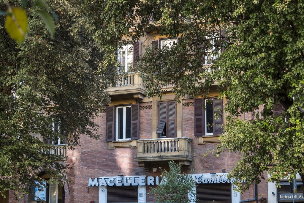A neighborhood macelleria below a Roman apartment building with brick walls, balconies, shutters, and trees framing the façade.