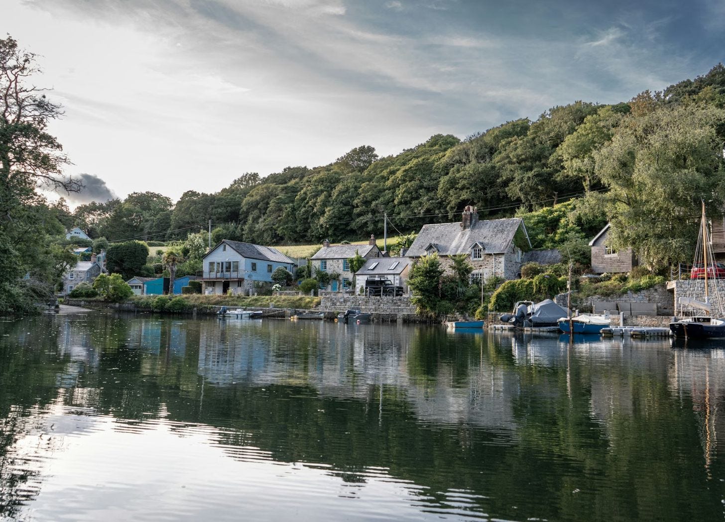 Small harbor village in Cornwall, England, with boats docked along calm water and homes lining the shoreline.