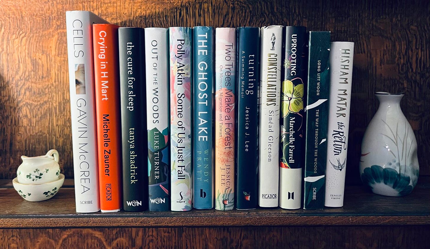 On a wooden shelf surrounded by two ceramic pots are hardback books shelved. There is Cells by Gavin McCrea, Crying in H Mart by Michelle Zauner, The Cure for Sleep by Tanya Shendrick, Out of the Woods by Luke Turner, Some of Us Just Fall by Polly Atkin, The Ghost Lake by Wendy Pratt, Two Trees Make a Forest by Jessica J. Lee, Turning by Jessica J. Lee, Constellations by Sinead Gleeson, Uprooting by Marchelle Farrell, The Way Through The Woods byLong Litt Woon, and The Return by Hisham Matar