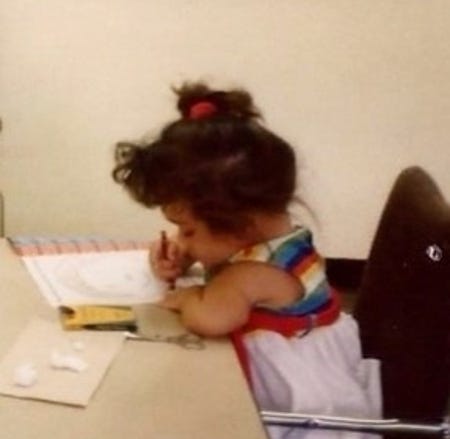 Gaelynn is four years old, sitting at a desk and coloring. She is wearing a white sundress that has rainbow stripes at the top and her hair is pulled into a messy bun.