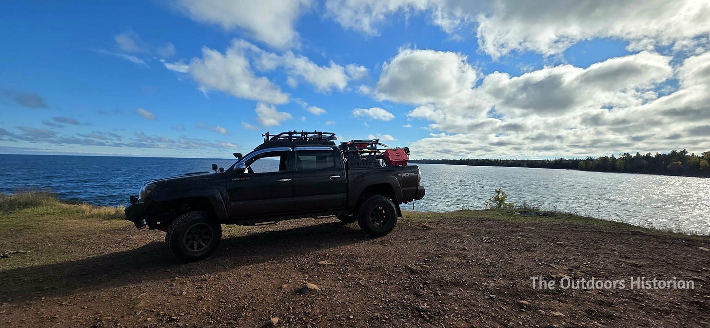 A side view of a black second generation Tacoma with roof racks and back racks on a gravel point in front of a blue Lake Superior and blue skies with some small white clouds.