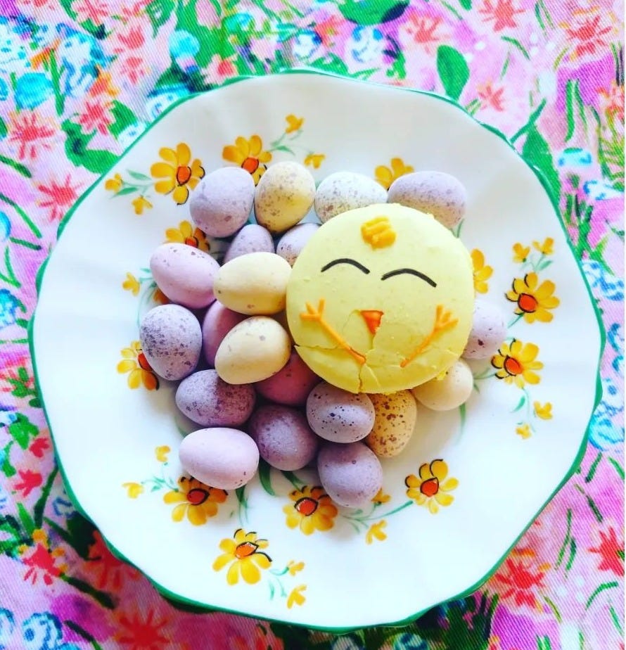A blue, pink and green floral table covering. On top is a green-edged white china saucer with yellow daisies filled with sugared mini-eggs in pastel pinks, lilacs and yellows. There is one lemon-yellow macaron, round with the iced face of a baby chick on it. The whole thing is a riot of spring tweeness but it is pretty too.