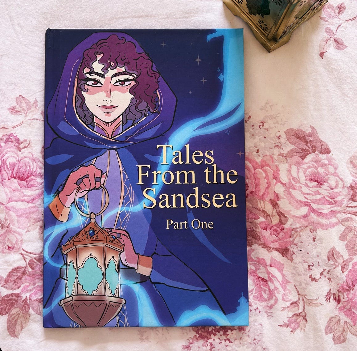 A photograph of The Tales of the Sandsea, an illustrated collection of interludes from THE STARDUST THIEF, set on a floral blanket. The cover features Loulie holding up a flaming blue lantern. (Art by Samohsai)