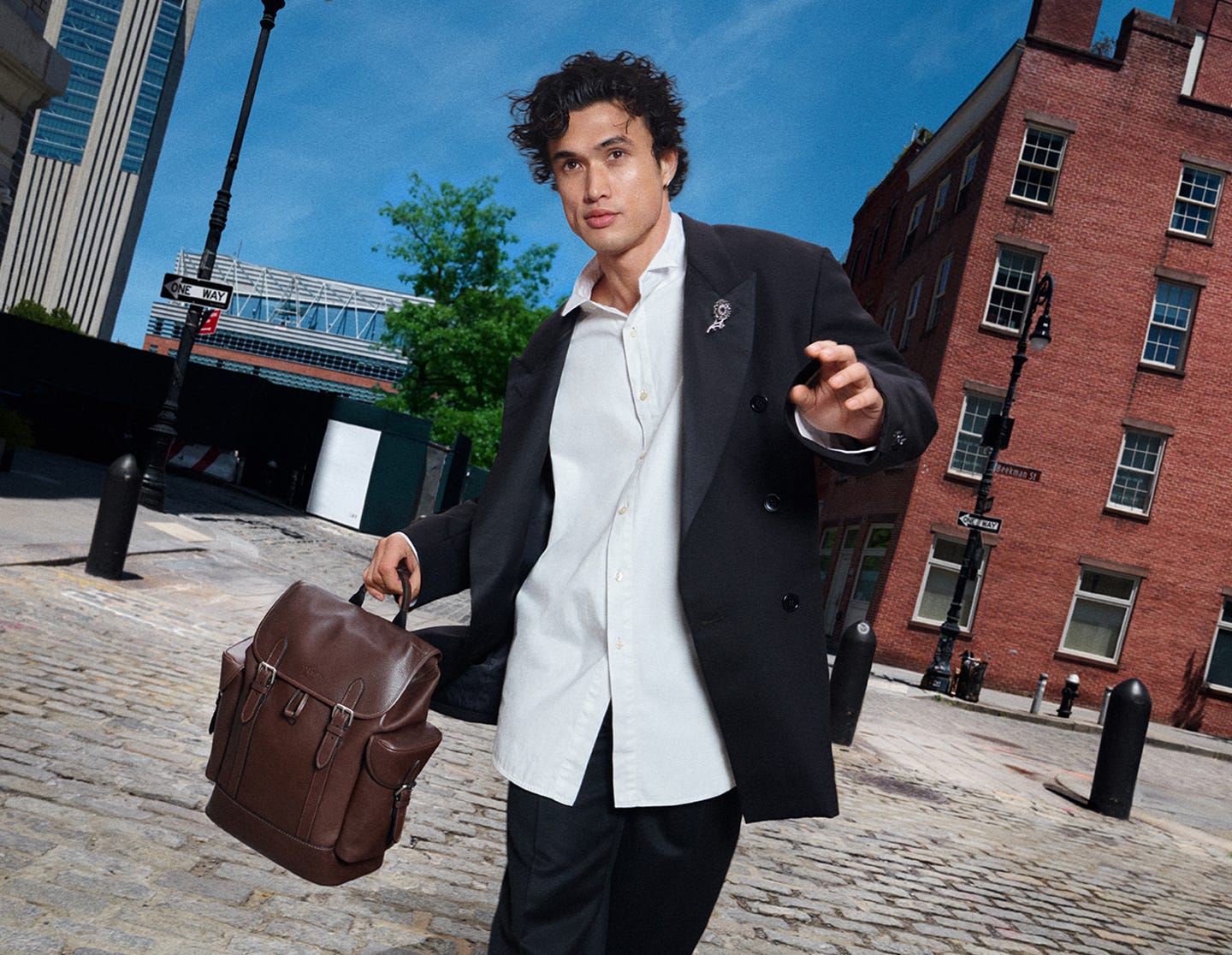 Young man in black blazer walking an urban cobblestone street carrying a brown leather backpack