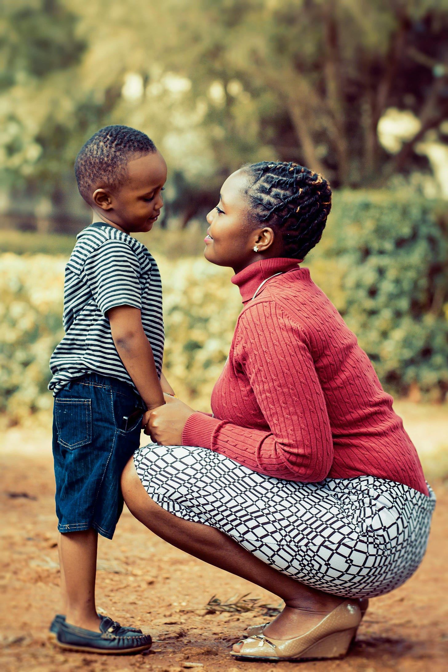 A young woman with braids close to her head, wearing a coral turtleneck sweater and black and white patterned skirt, squats down in front of a child, maybe 4 or 5, and holds his hands. She is smiling at him and her eyes are soft and half closed. He is looking at her with his head tilted and eyes lowered; he seems shy or perhaps like he was recently upset, but he also looks relaxed and comforted by her presence. A young woman with braids close to her head, wearing a coral turtleneck sweater and black and white patterned skirt, squats down in front of a child, maybe 4 or 5, and holds his hands. She is smiling at him and her eyes are soft and half closed. He is looking at her with his head tilted and eyes lowered; he seems shy or perhaps like he was recently upset, but he also looks relaxed and comforted by her presence.