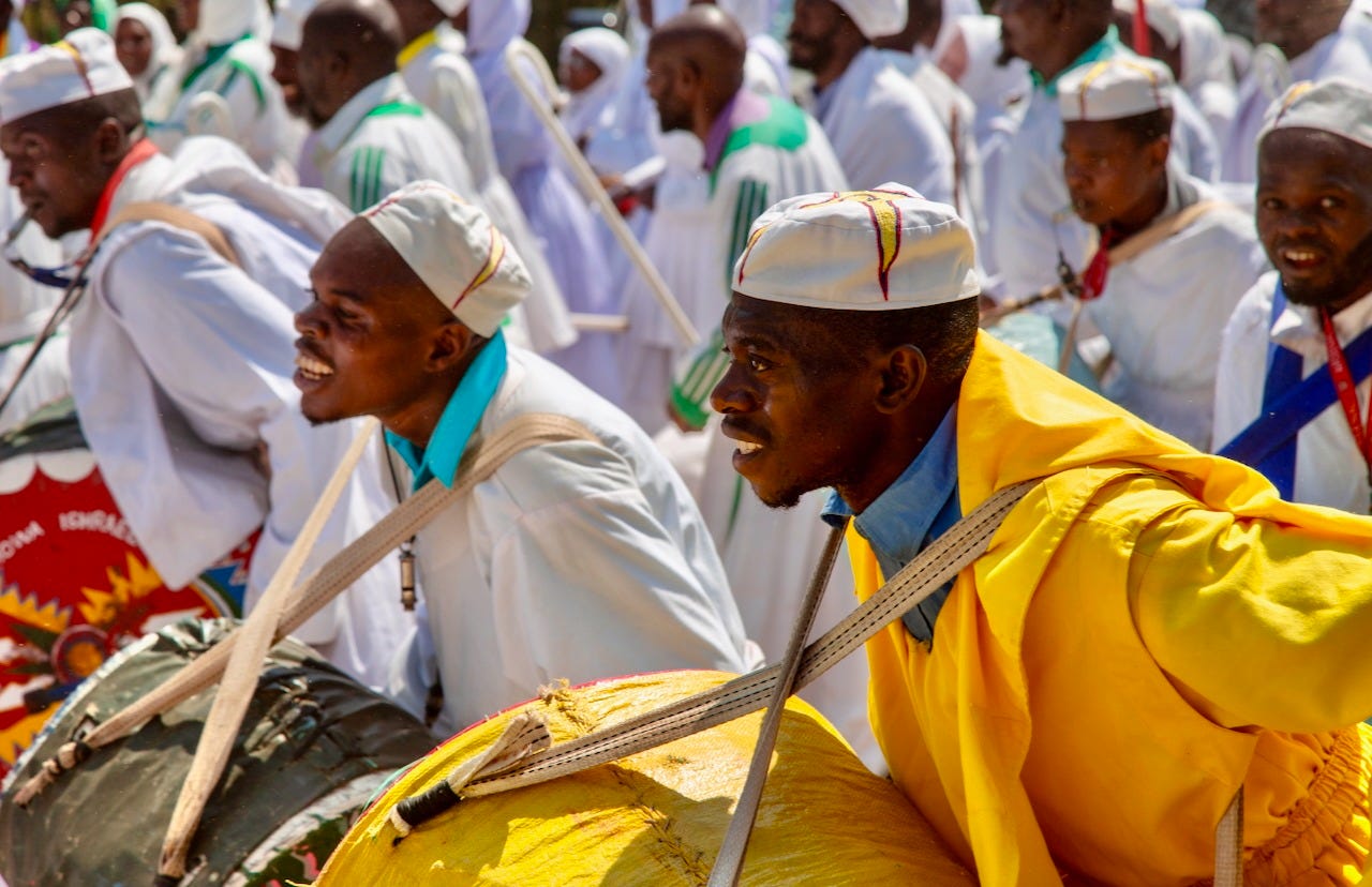 Drummers in white caps and robes, one in a bright yellow cloak, lean forward mid-procession with large drums slung across their chests, a crowd in white behind them. Drummers in white caps and robes, one in a bright yellow cloak, lean forward mid-procession with large drums slung across their chests, a crowd in white behind them.
