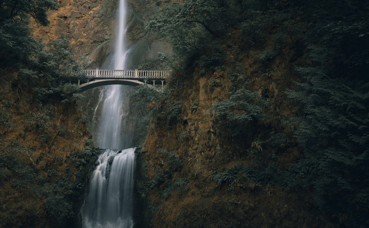 photo of waterfall tumbling down a steep brown and green hill with a gray stone bridge about halfway up the falls