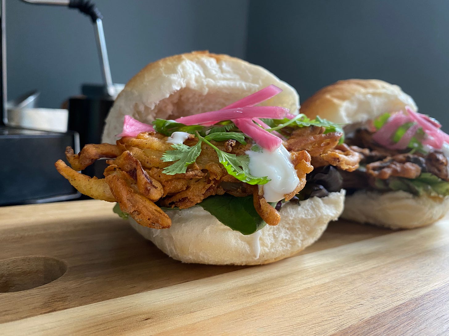 Two bread rolls, filled with salad leaves, fried onion bhaji, pink onion slices and yoghurt. Rolls are served on a wooden board.
