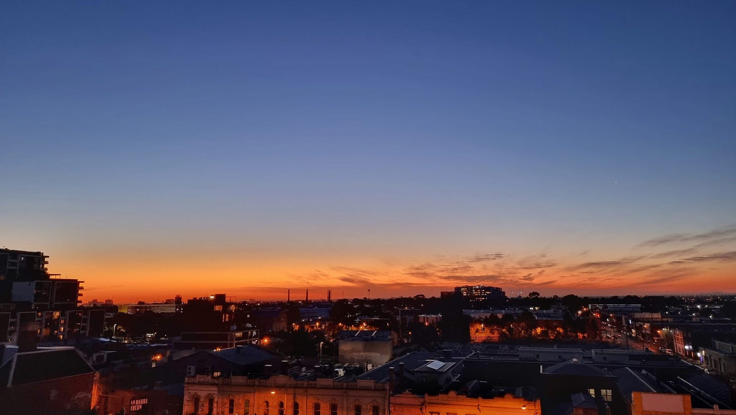 A large and empty sky that fades from dark blue to a red-orange where these tones are echoed in the building roof-tops and street-light-lit façades. 