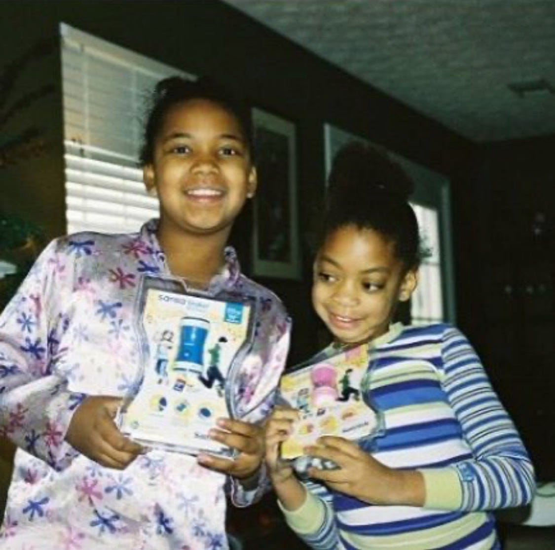 A photo of two young children on Christmas morning. The child on the left (the author of this piece) is wearing a set of pink satin pajamas with purple and pink six-pronged figures patterened on it. The child on the right is the author's sibling who wears a green, white, and purple striped sweater. Both children are holding Sansa Shakers, MP3 players that they both received as gifts. 
