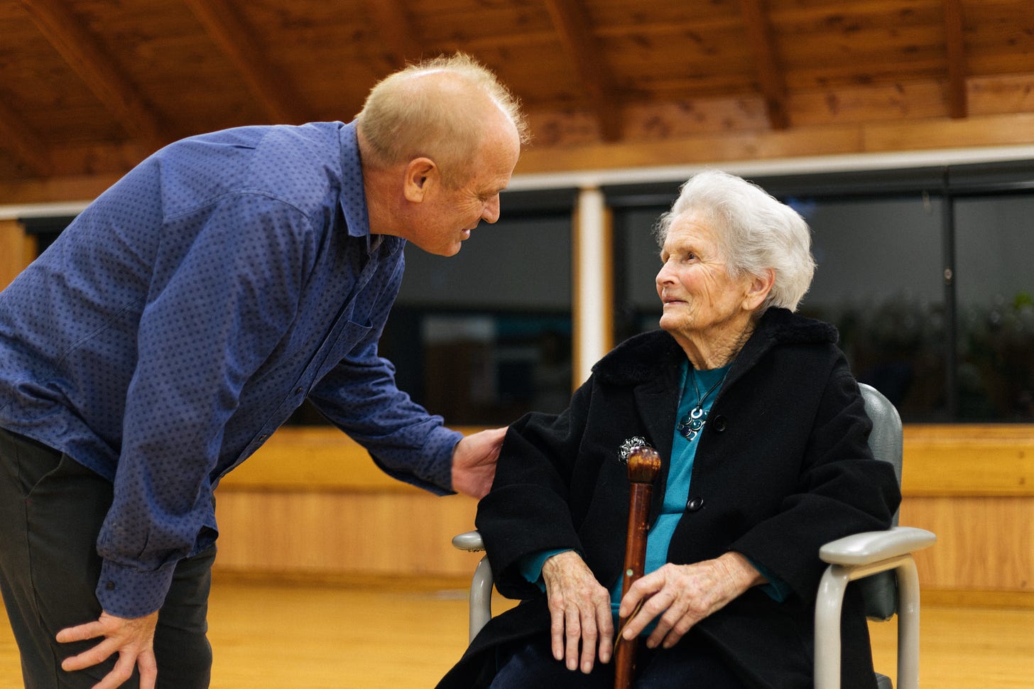 A photograph: my uncle David is on the left, he's wearing a blue shirt with small black dots on it, and is leaning down and holding the arm of my grandmother, on the right, who is sitting in a chair and holding a walking stick, and looking up at him.
