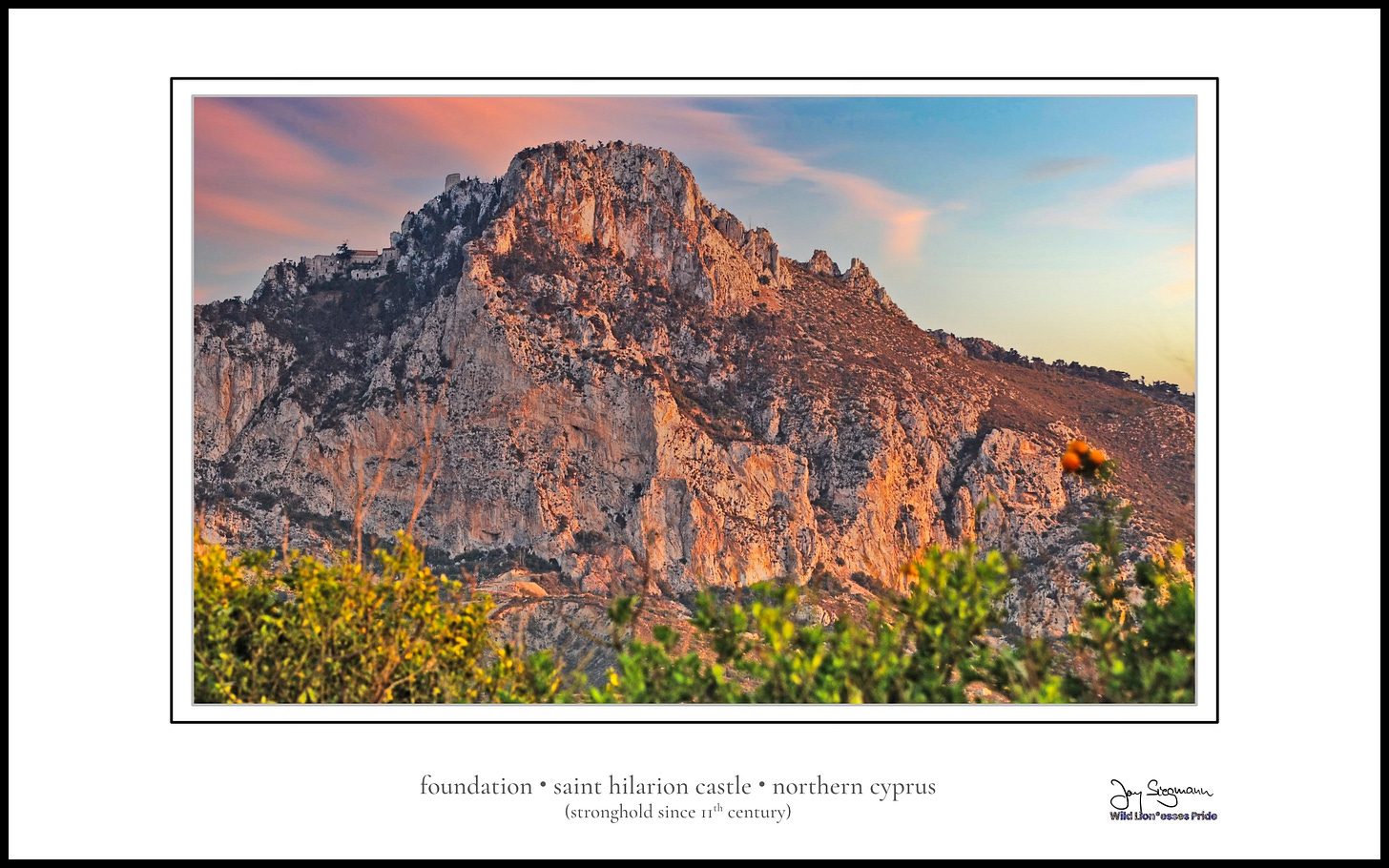 Saint Hilarion Castle in northern Cyprus, perched atop a rugged mountain peak in the Kyrenia range. The castle’s stone walls and towers crown the rocky summit, bathed in warm sunset light. Foreground shrubs blur into view, while the sky glows with soft pink and blue streaks.