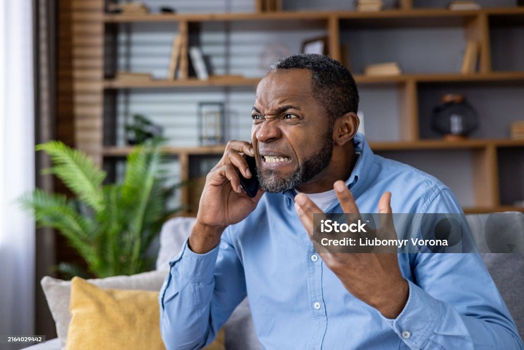 Frustrated man yelling on smartphone in living room with bookshelf background Angry man shouting on phone in living room expressing frustration. Person wearing blue shirt, sitting on gray couch with beige pillow, plant and bookshelf in background. 40-44 Years Stock Photo