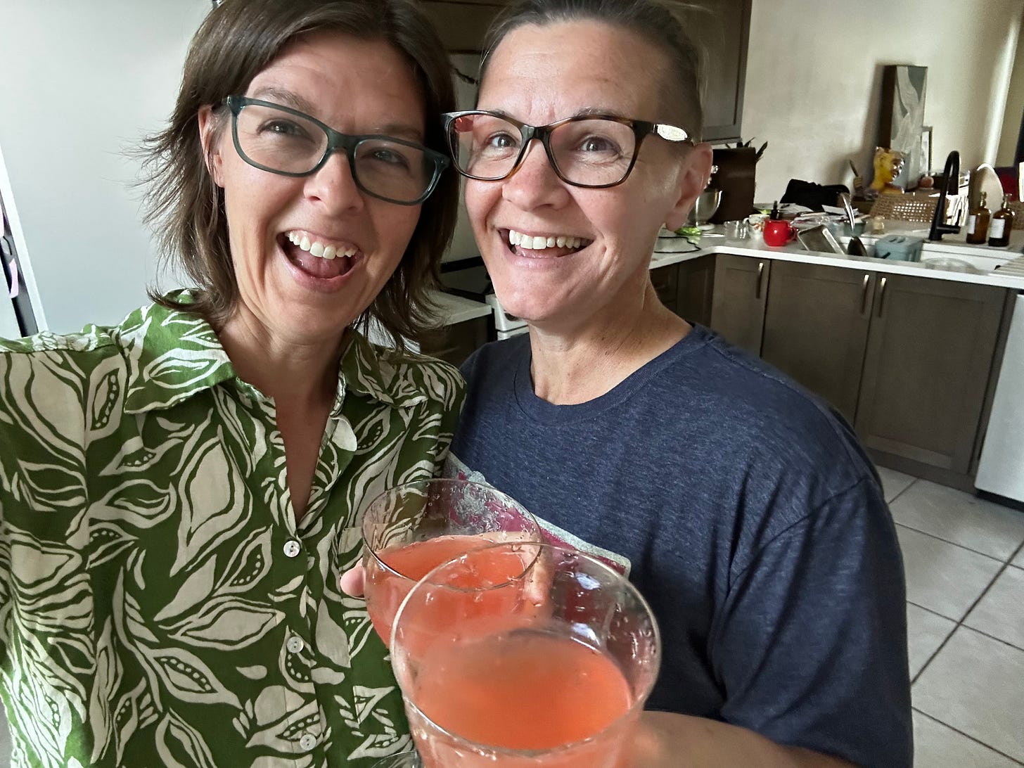 Two women wearing glasses, smile and cheers glasses of grapefruit juice in their kitchen.