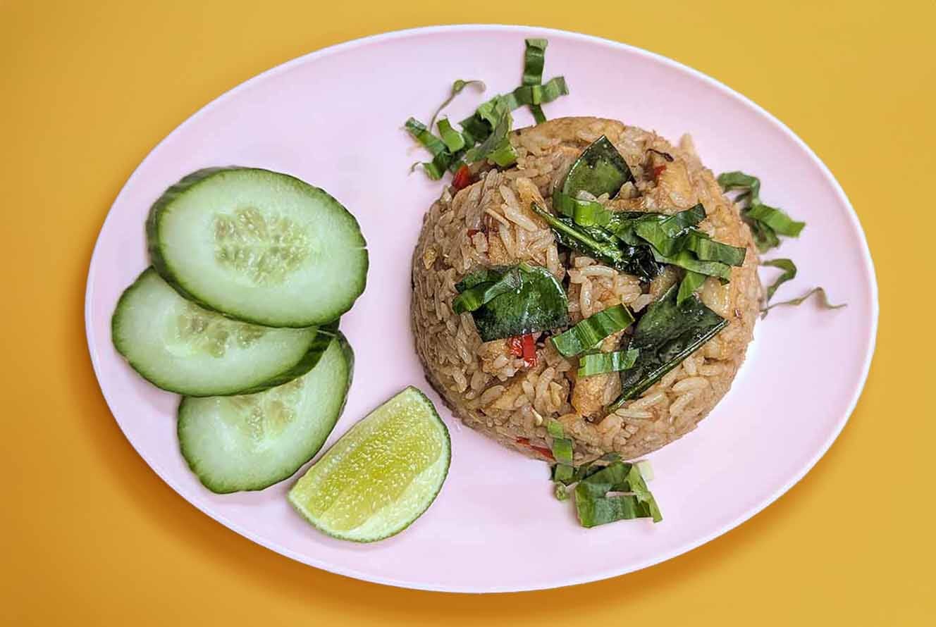 Top down view of a pink oval plastic plate with a moulded round of fried rice topped with chopped herbs. Alongside the rice are three slices of cucumber and one slice of fresh lime.