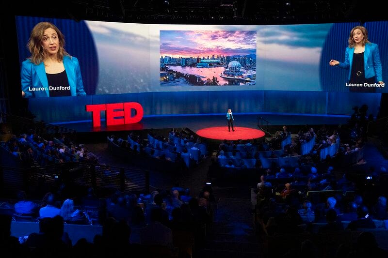 Lauren Dunford delivering a TED Talk on a large stage with a projected city image behind her. Lauren Dunford delivering a TED Talk on a large stage with a projected city image behind her.
