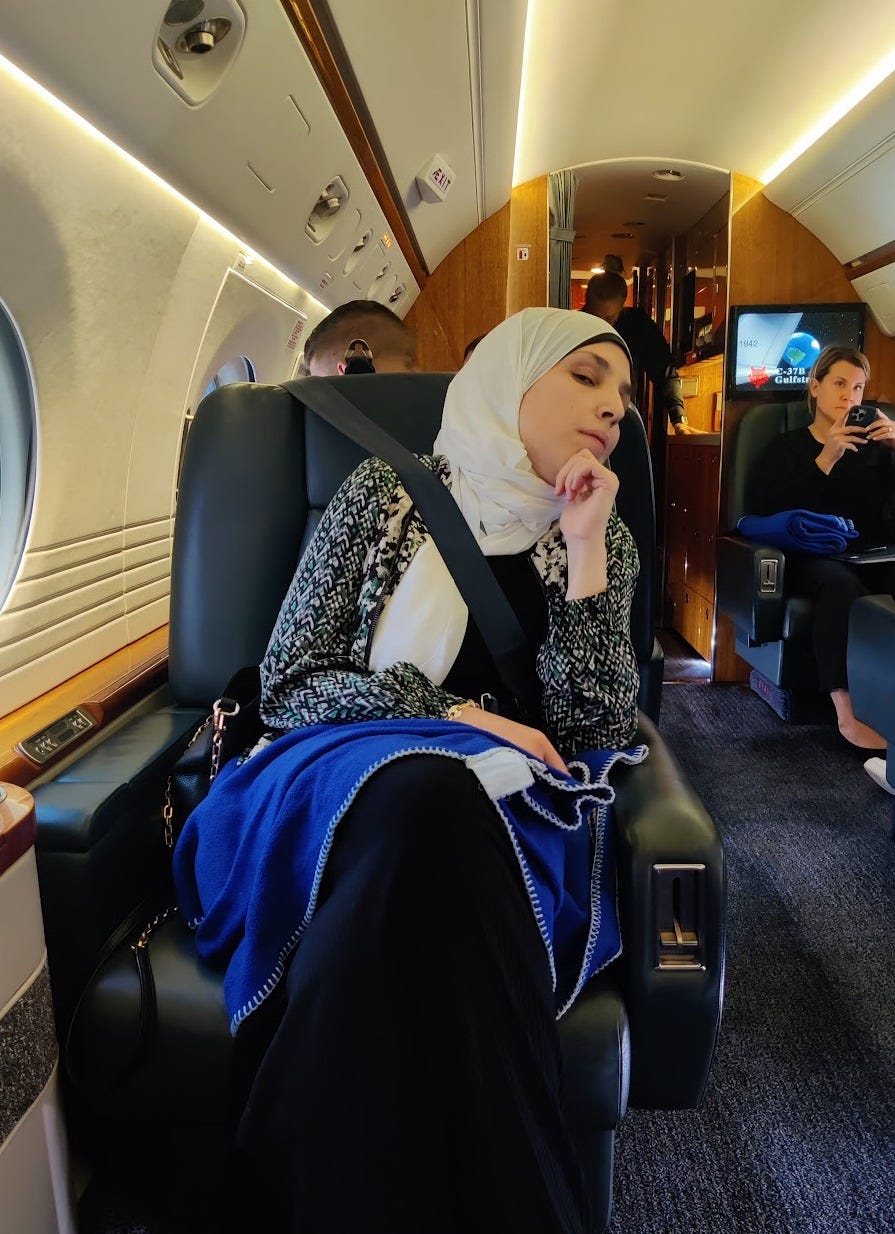 Sara sits in a spacious airplane seat. She wears a white hijab, white blouse, and a black and white patterned jacket. 
